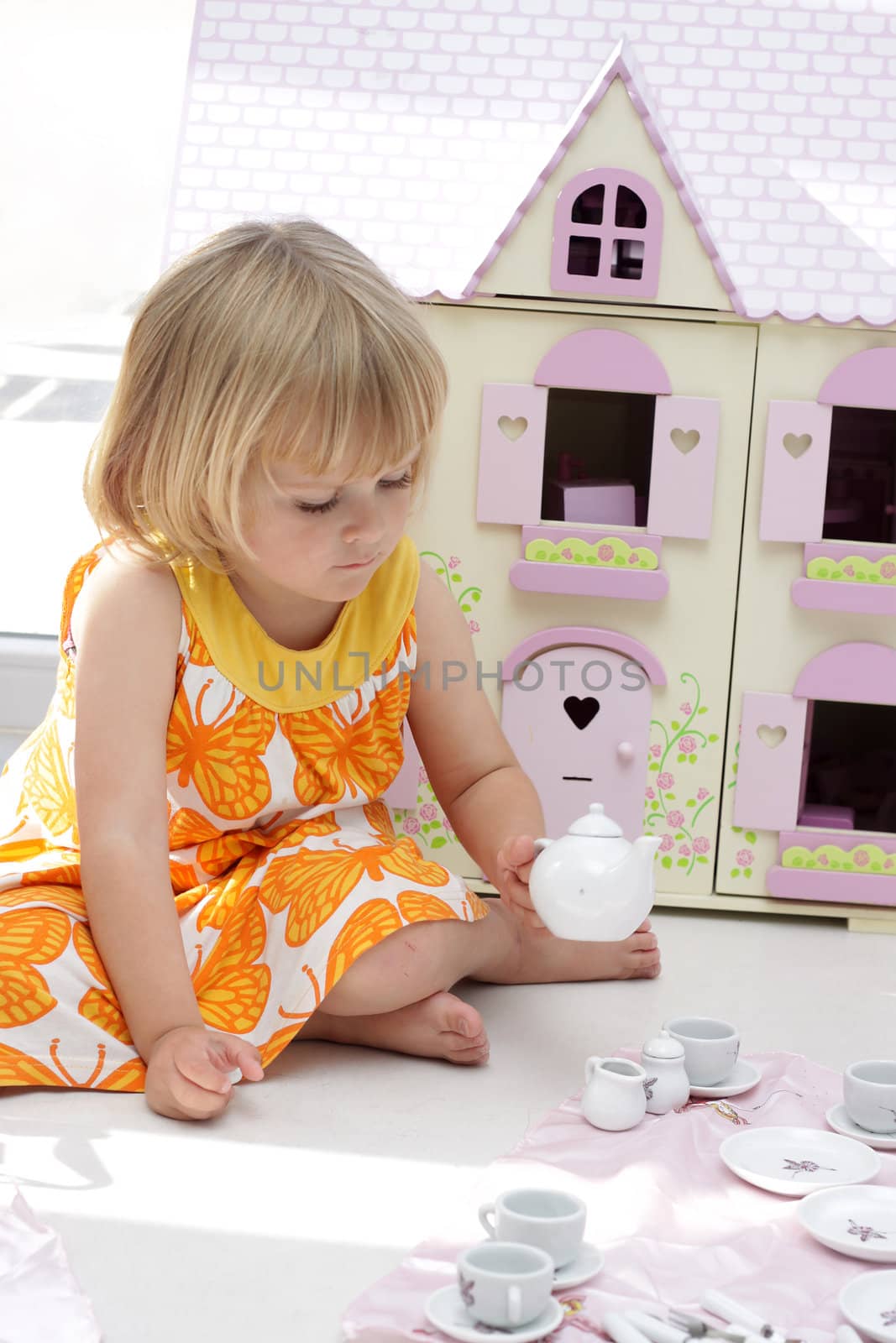 A little 4 year old girl playing tea party with her teapot set in front of a wooden dollhouse