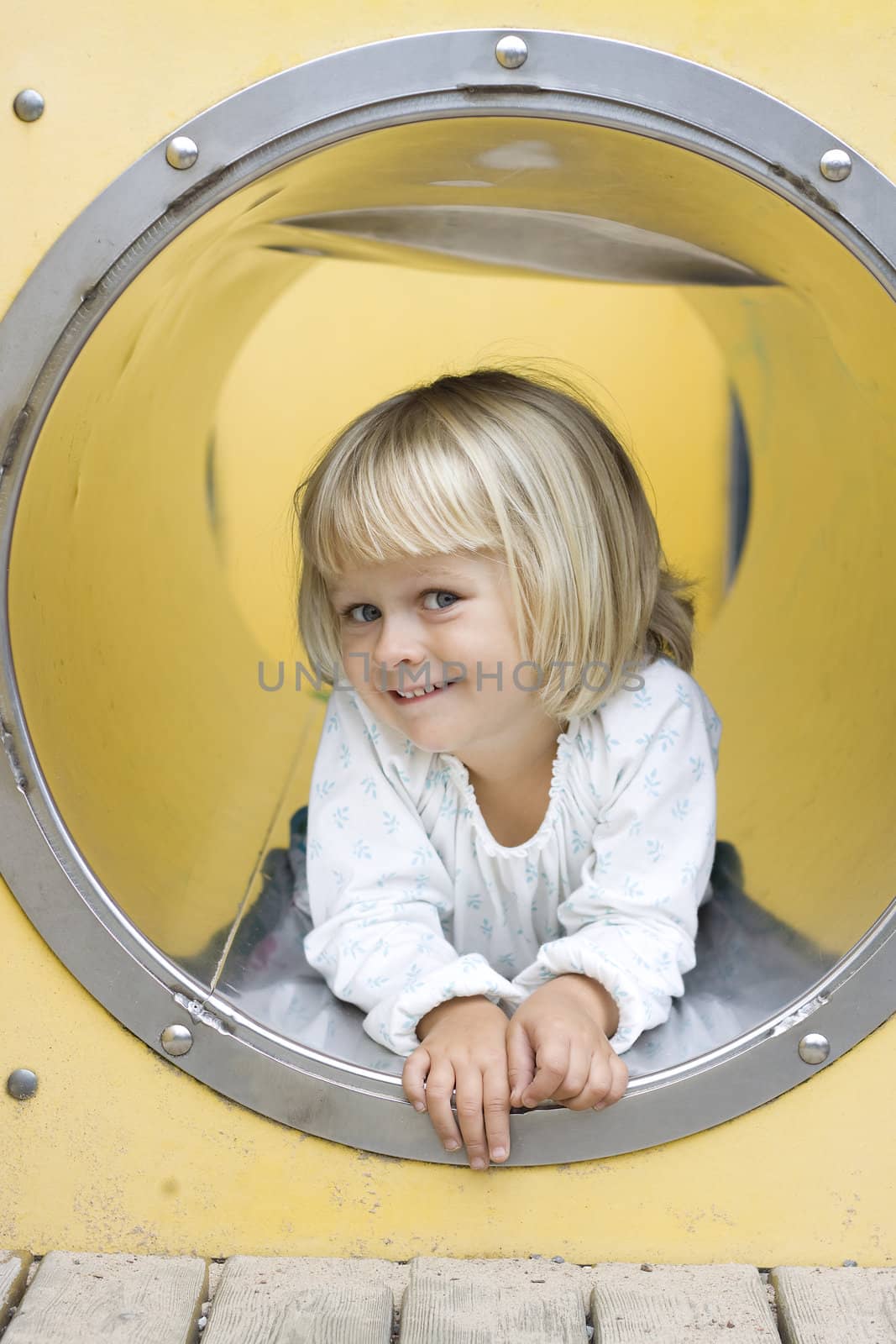 A cute little girl peeking out from a play ground tunnel, smiling at the camera. 