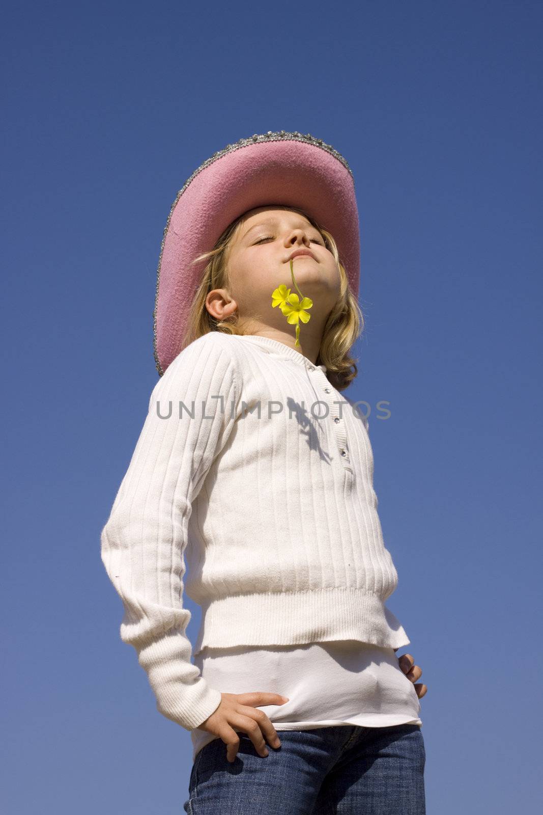 A young girl with hat and a flower in her mouth soaking up the sun. Closed eyes. Isolated on blue background. 