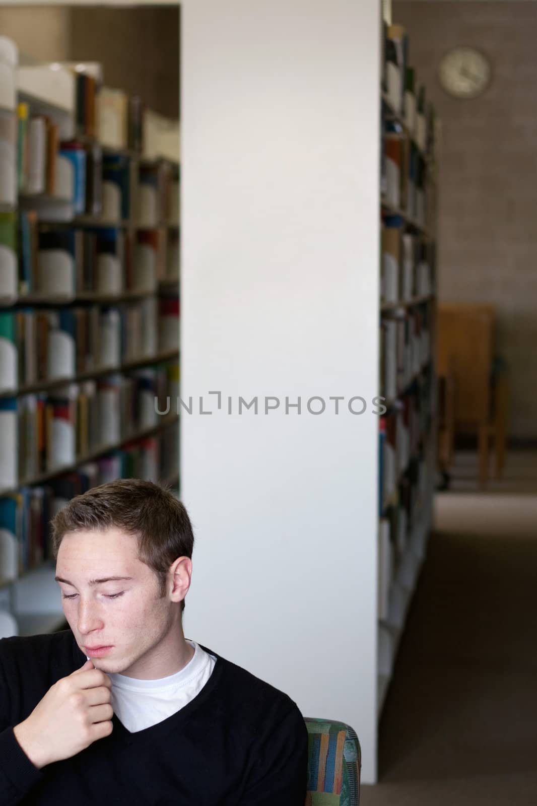 A young university student thinking about something or reading in the library with a wall clock visable in the background.