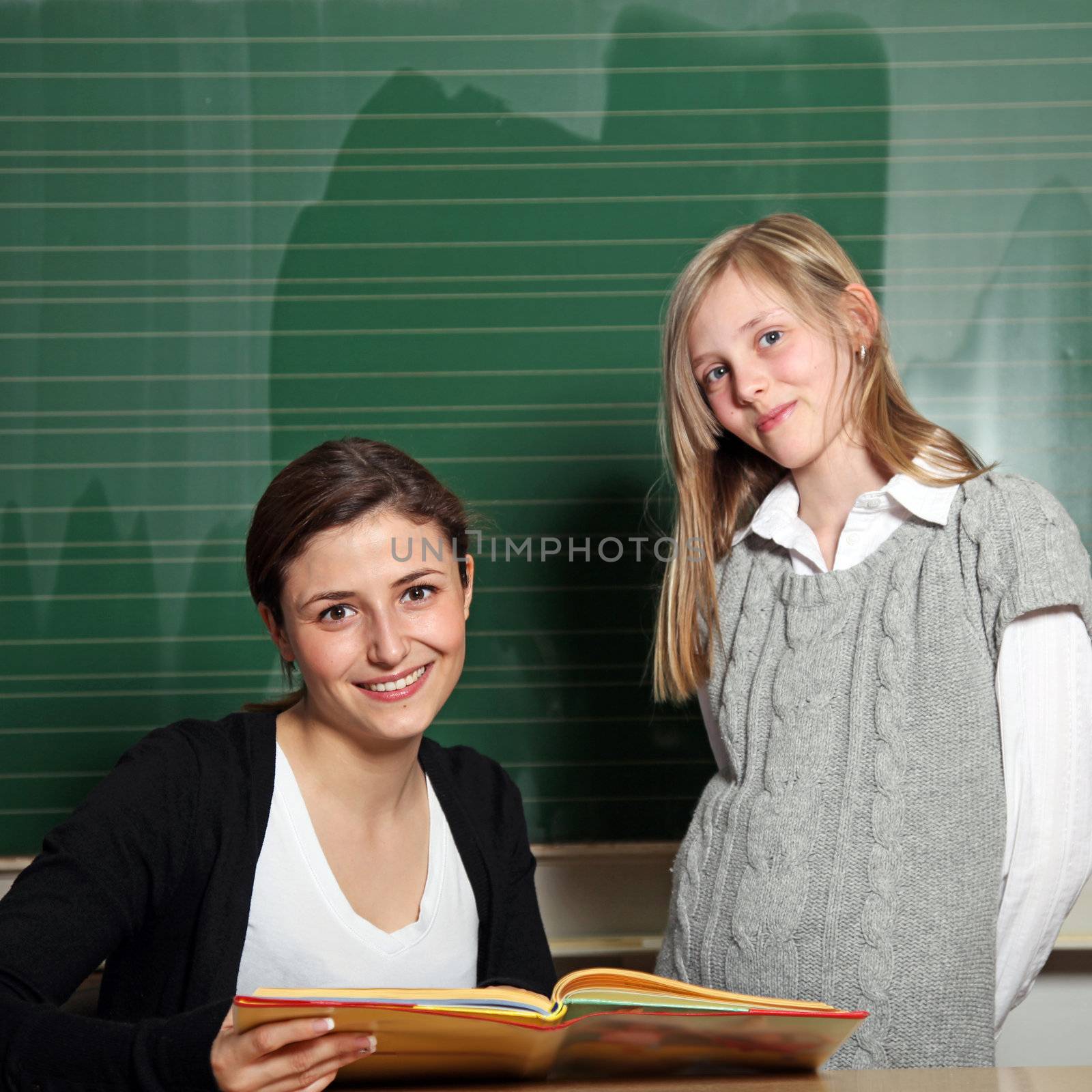 Nice teacher with a textbook and a student at the blackboard. They both smile at the camera