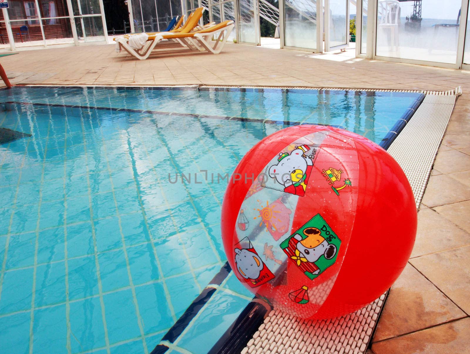 Swimming pool  scene: brihgt colored red inflatable ball close to the water.
Nice contrast between red and blue.  Clear reflection of glass wall on a  smooth water surface.