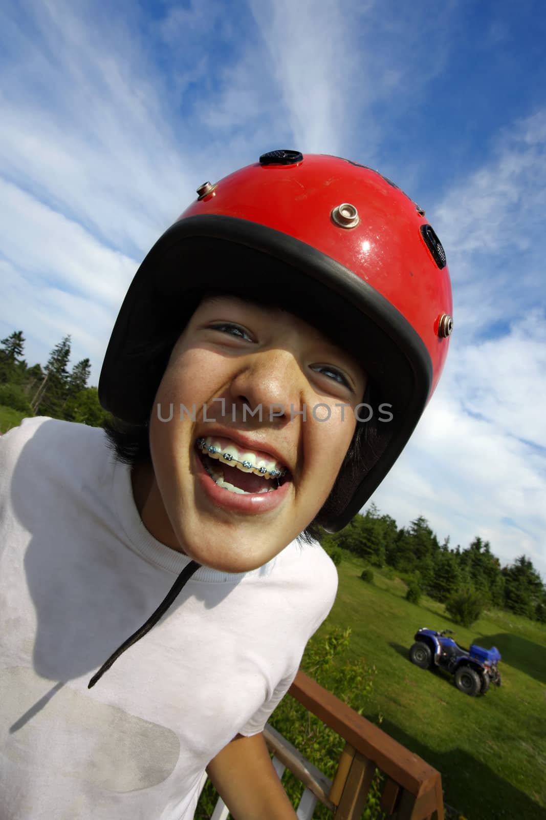 An ecstatic Asian boy with red helmet, getting ready to ride an all terrain vehicle parked in the background.
