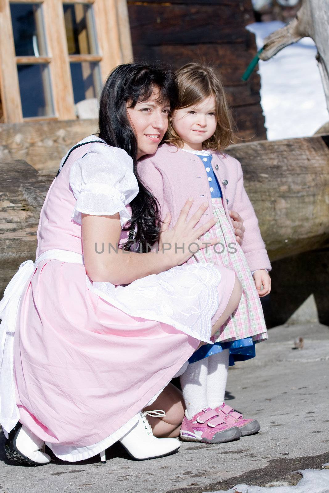 Mother and infant in costume stands in front of a cabin in the mountains