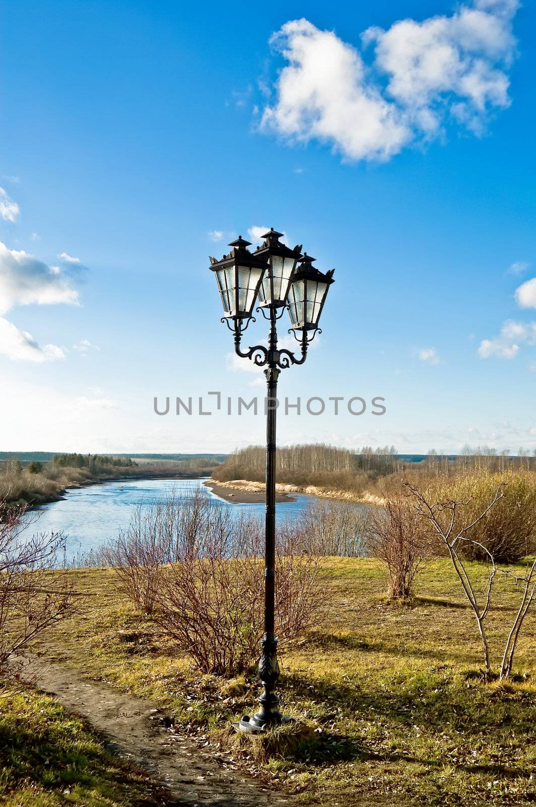 Black decorative lamp with three shades against the blue sky with clouds, paths, grass, bushes, trees and rivers