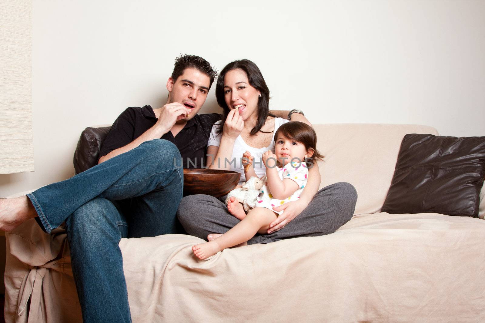 Beautiful happy family, father mother toddler daughter child, eating snacks while sitting on sofa couch in livingroom.