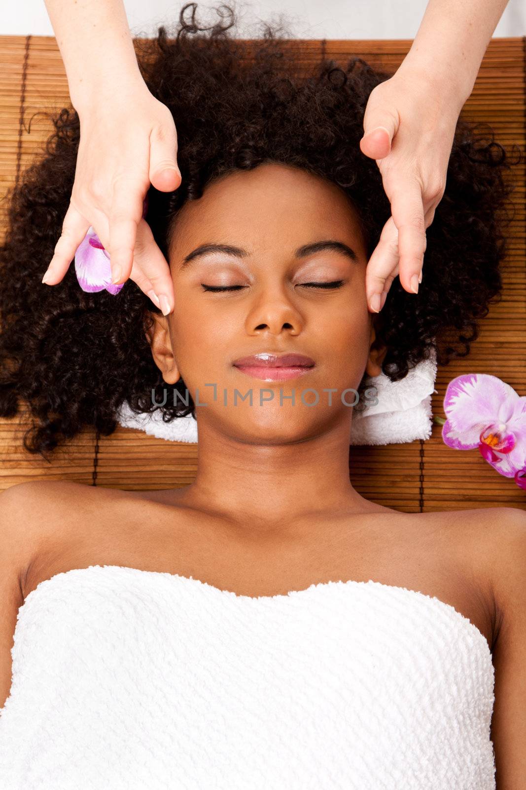 Beautiful happy peaceful sleeping woman at day spa, laying on bamboo table with head on pillow wearing a towel getting a facial temple massage, isolated.