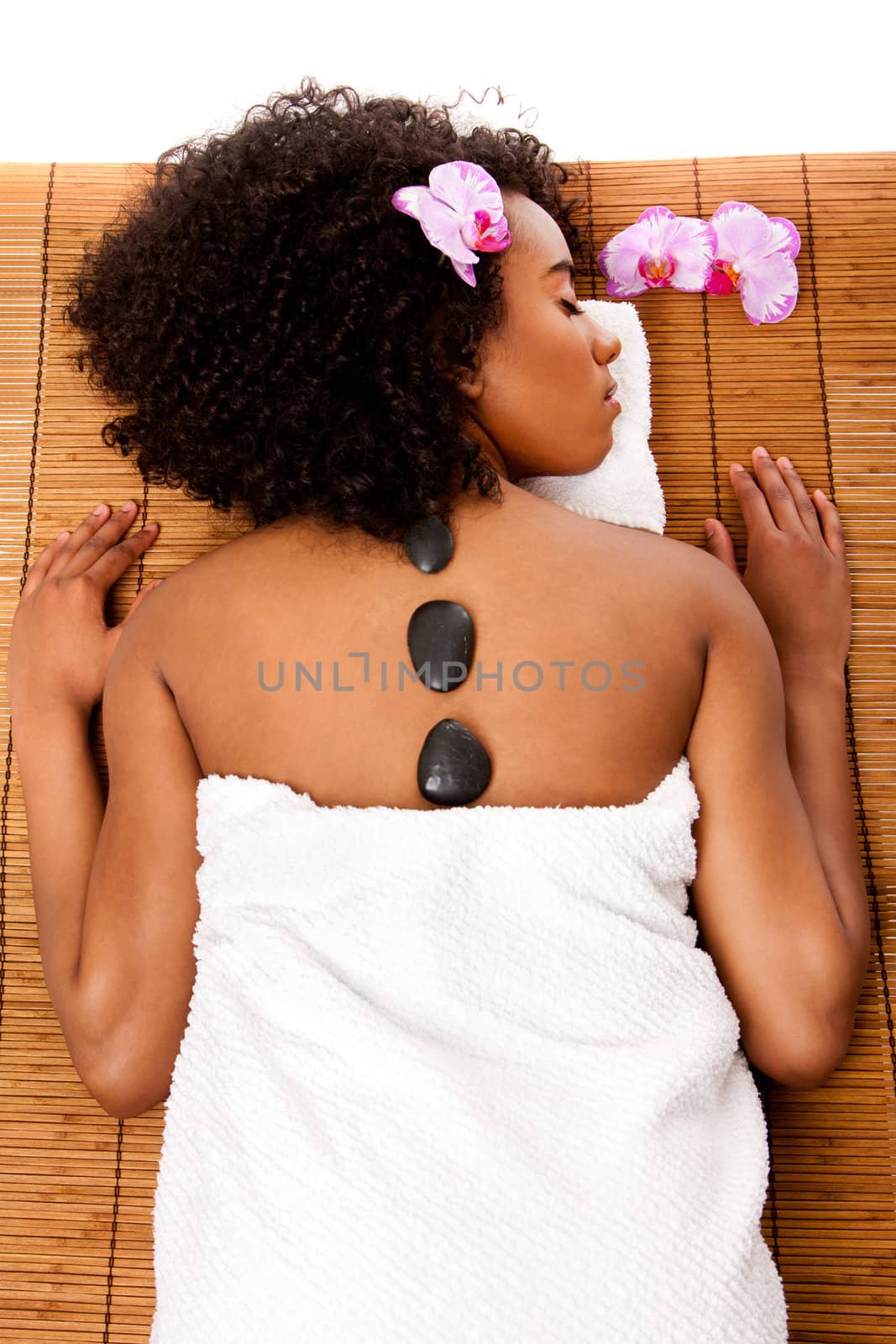 Beautiful happy relaxed Latina-African woman at health day spa with hot lastone therapy massage treatment laying in white towel on bamboo table decorated with orchids.