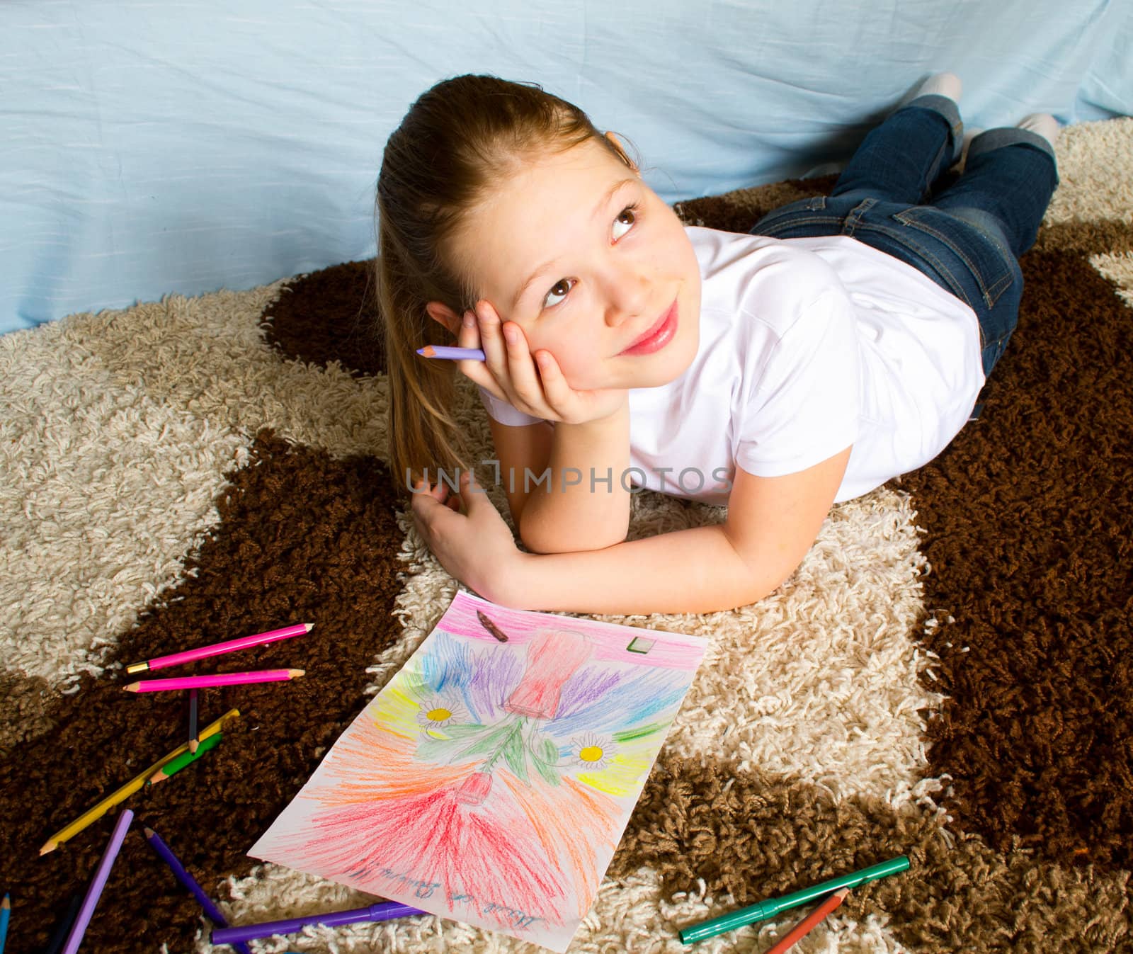 The girl lies on a carpet and thoughtfully draws color pencils a picture