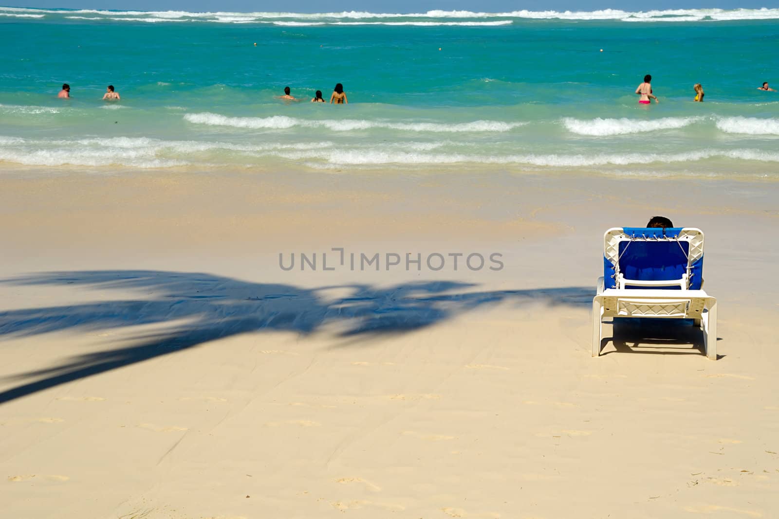 Woman is resting in a sunbed infront of sea. In the bacground people are bathing in the water. 