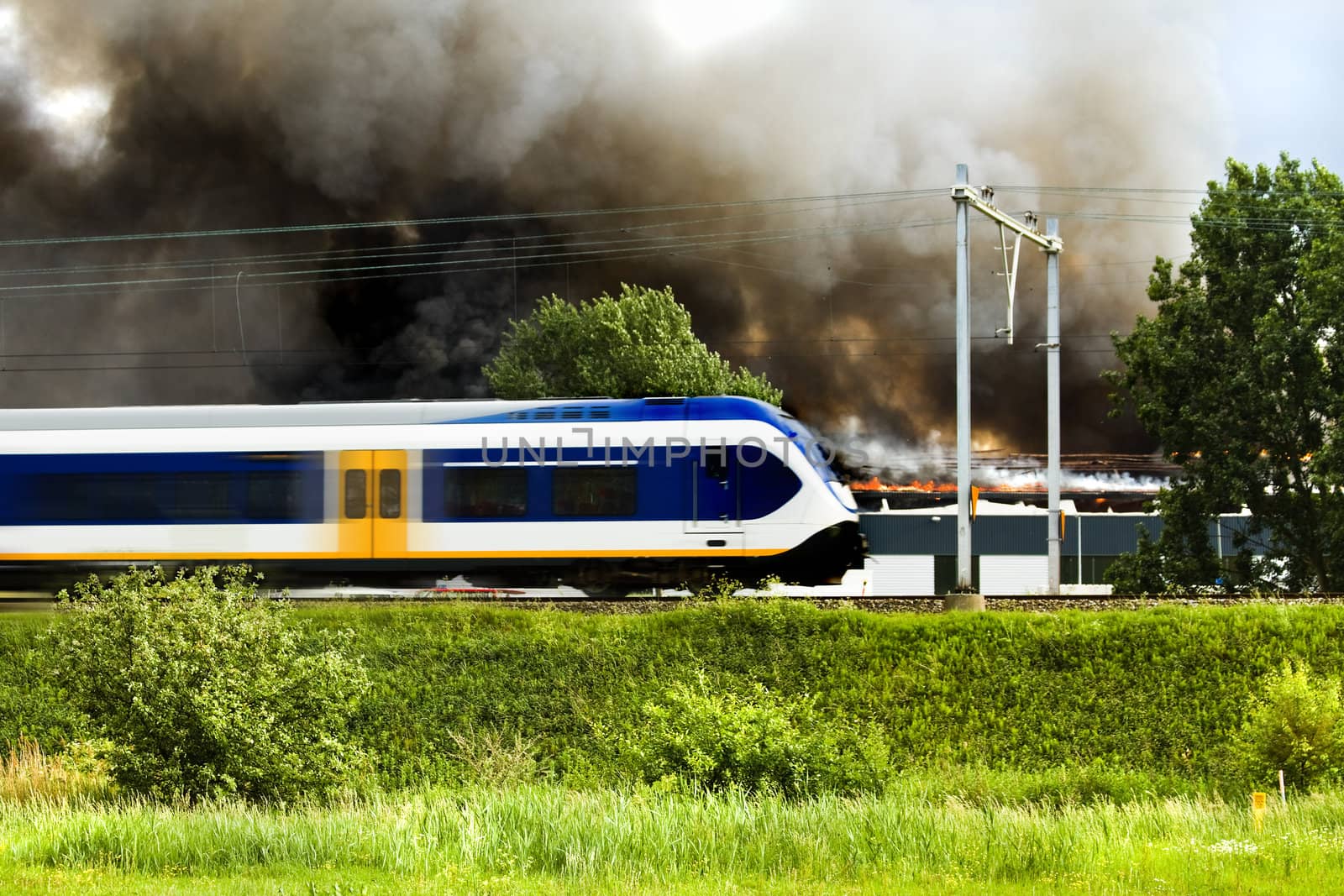 Train passes by on railway embankment while in the background a factory is burning to the ground