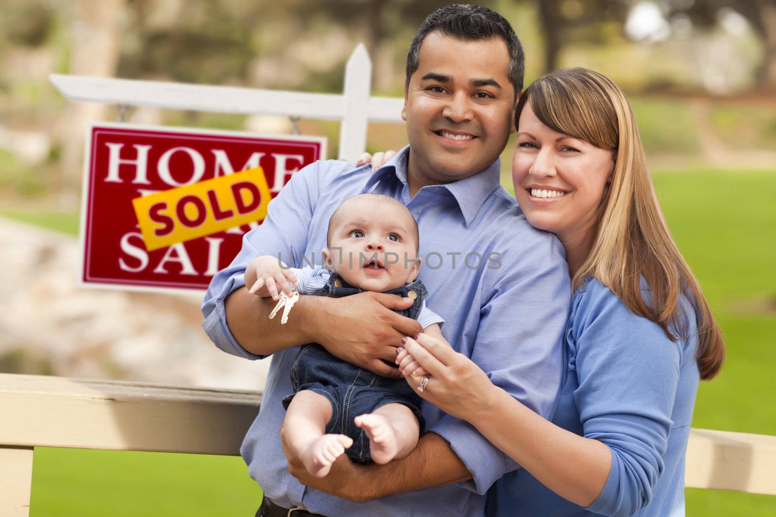 Happy Mixed Race Couple with Baby in Front of Sold Real Estate Sign.