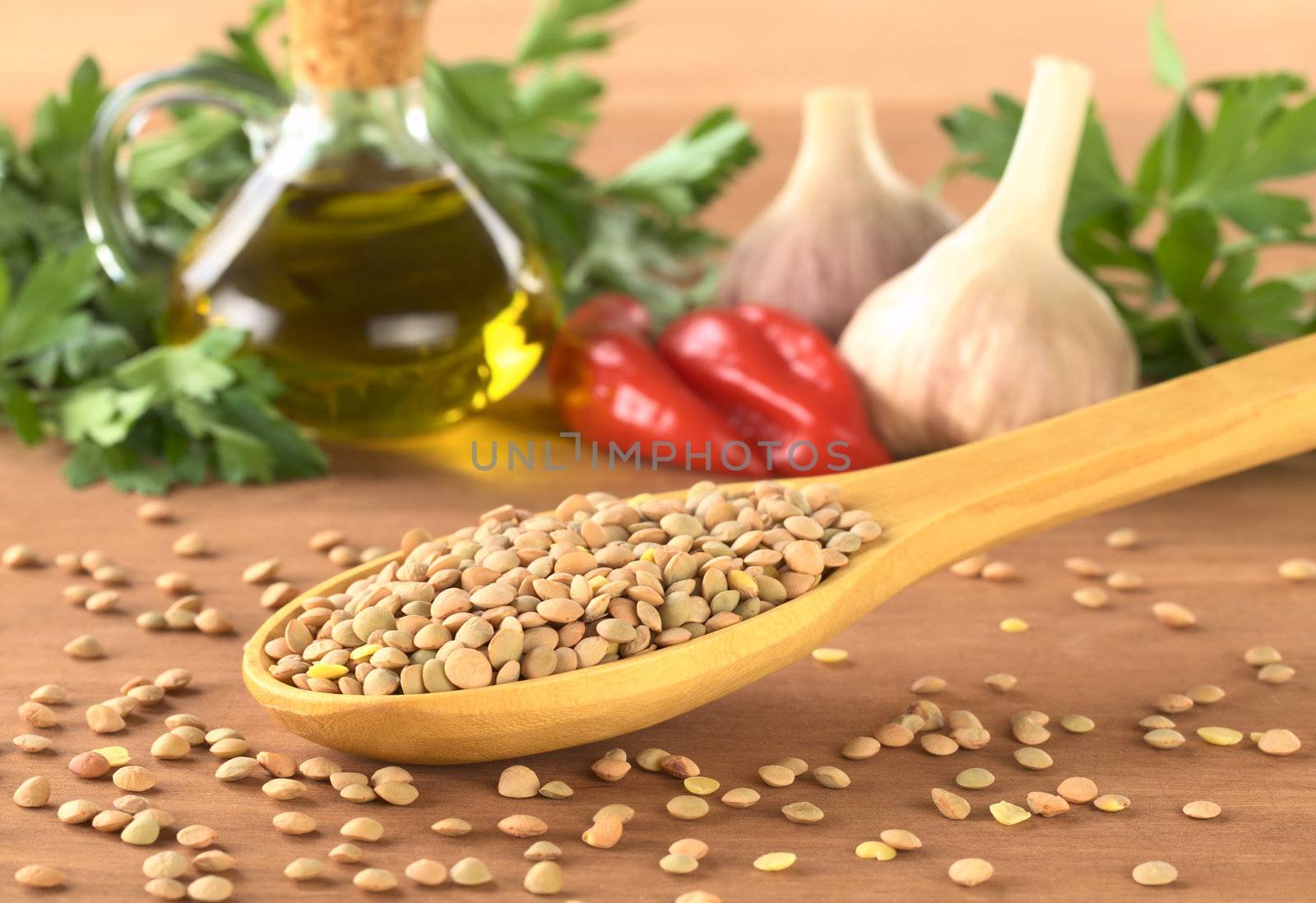 Raw lentils (lat. Lens culinaris) on wooden spoon with condiments in the back such as oil, hot pepper, garlic and parsley (Selective Focus, Focus on the lentils in the first half of the spoon)