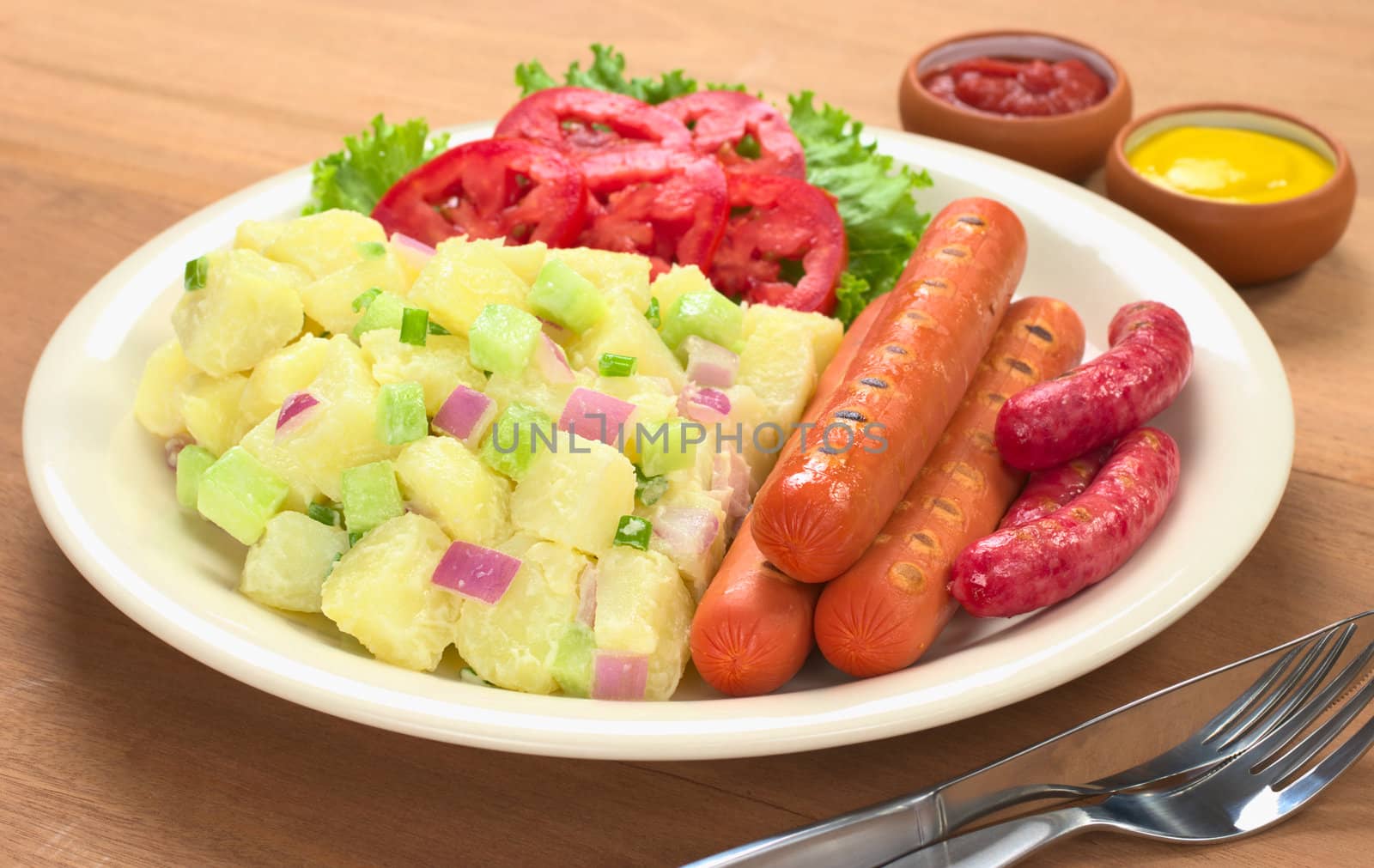 Sausages and potato salad with red and green onions and cucumber with a mayonnaise-cream dressing and tomato slices on plate with ketchup and mustard in the back (Selective Focus, Focus on the front of the meal)