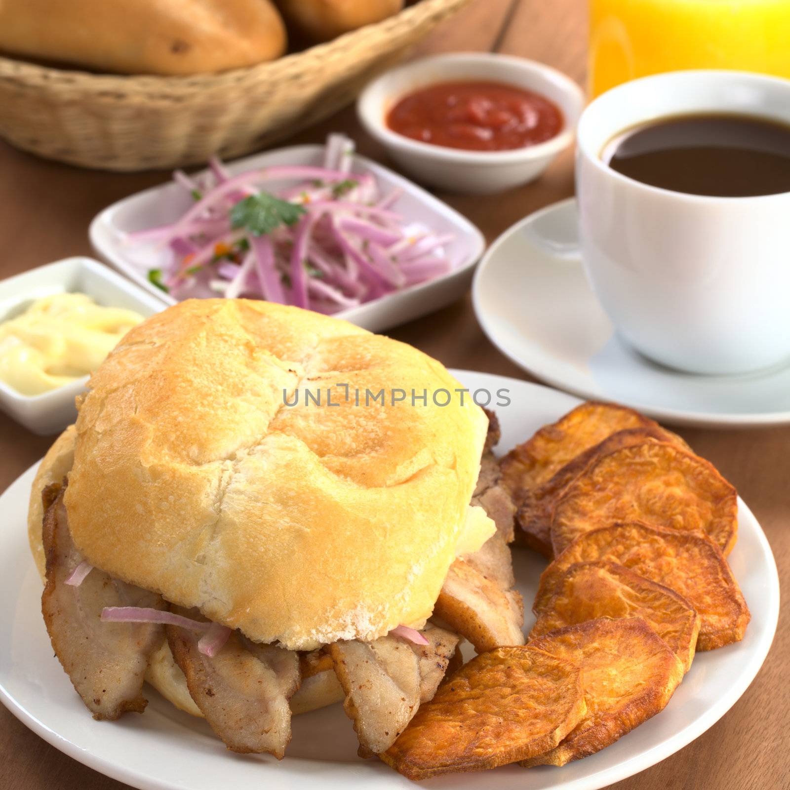 Typical Peruvian breakfast consisting of Pan con Chicharron (Bun with fried meat) and fried sweet potato, salsa criolla (onion salad), ketchup, mayonnaise, with coffee, orange juice and buns (Selective Focus, Focus on the front of the bun and the meat)