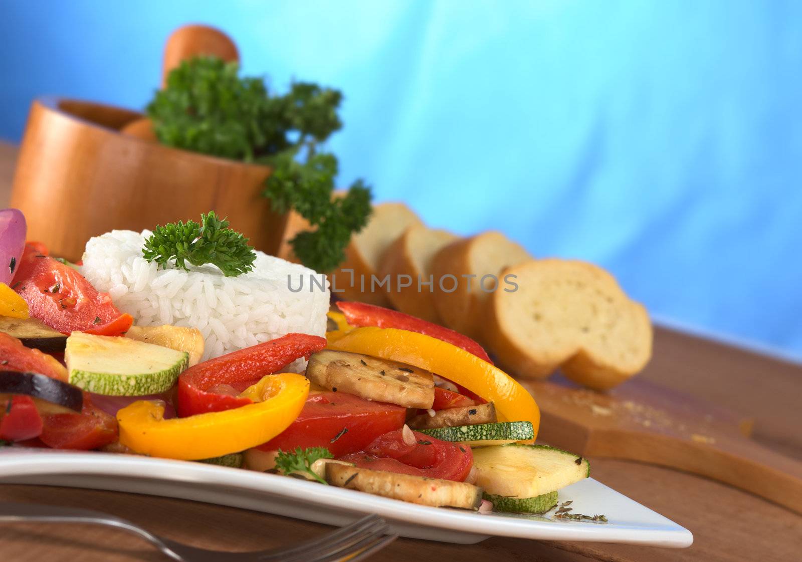 Ratatouille of zucchini, eggplant, tomato, bell pepper and onion with cooked rice and mortar with herbs and baguette in the back (Selective Focus, Focus on the front of the rice, the parsley on top and the vegetable around)