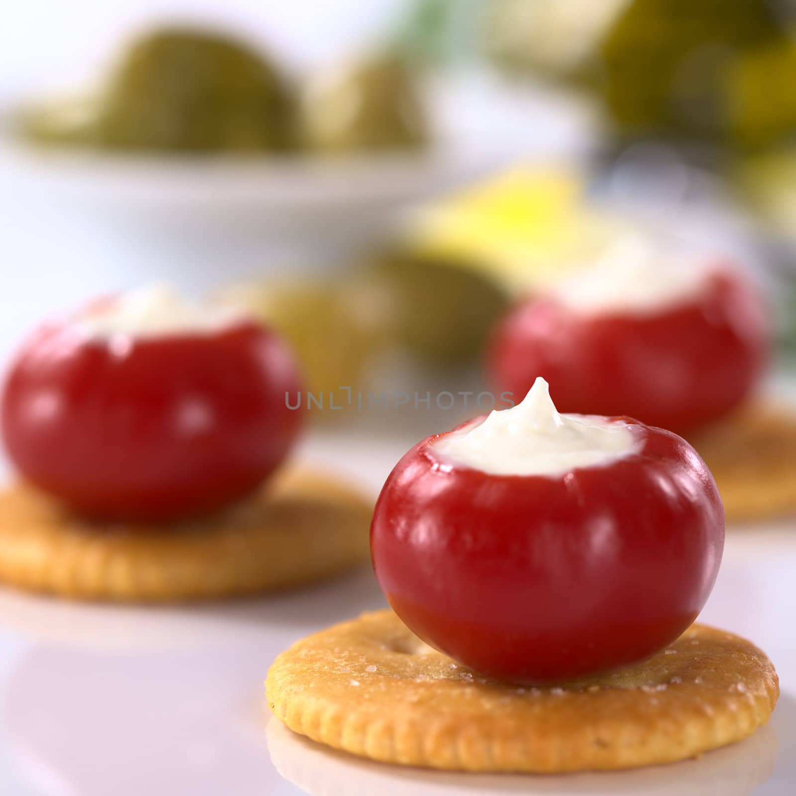 Small round hot red pepper filled with cream cheese on salty crackers with green olives in the back (Very Shallow Depth of Field, Focus on the tip of the cream cheese filling in the first pepper)