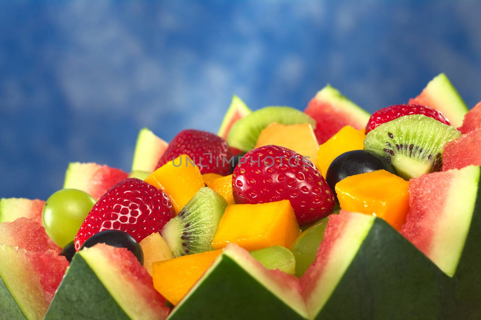 Fresh fruit salad of mango, strawberry, kiwi and grapes in a bowl from watermelon in front of blue background (Selective Focus, Focus in the middle of the fruit salad) 