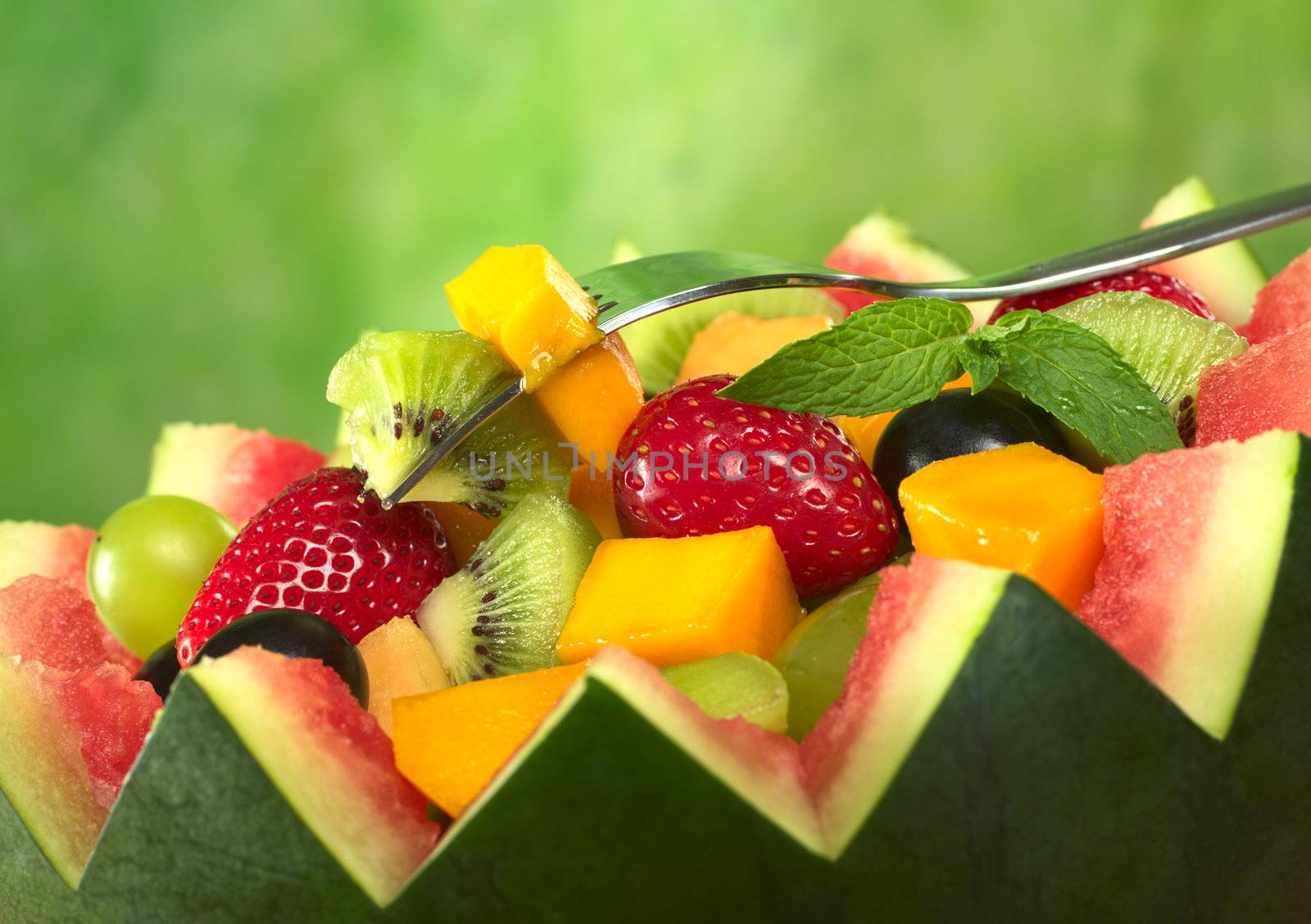Fresh fruit salad (strawberry, kiwi, mango, grape) in melon bowl with kiwi and mango on fork and a mint leaf as garnish in front of green background (Selective Focus, Focus on the fruit on the fork and the mint leaf)
