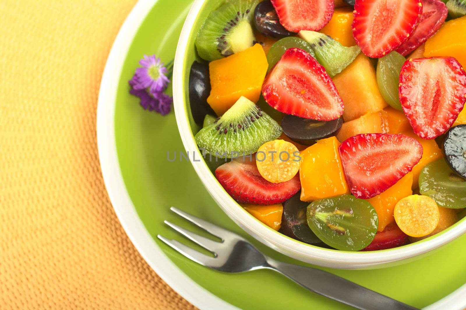 Fresh and healthy fruit salad with strawberry, kiwi, grape, mango and physalis in a bowl with fork and small blue flowers photographed from above (Selective Focus, Focus from the front to the middle of the bowl)