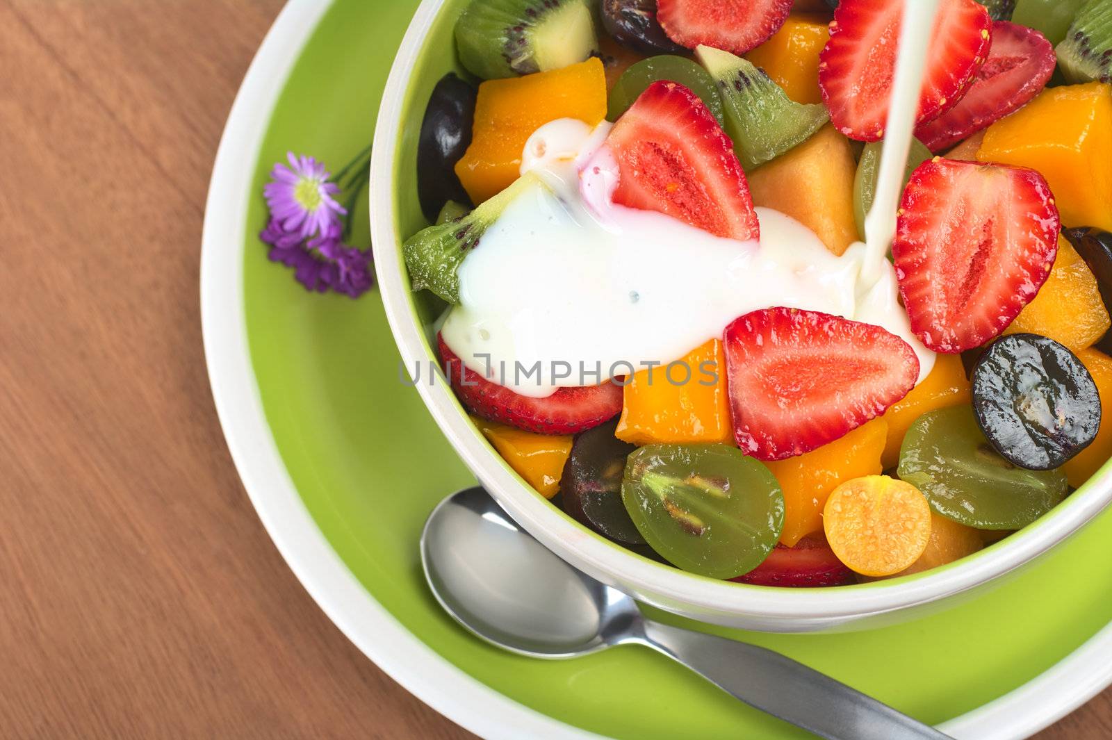 Fresh and healthy fruit salad with strawberry, kiwi, grape, mango and physalis in a bowl with plain yoghurt being poured over 