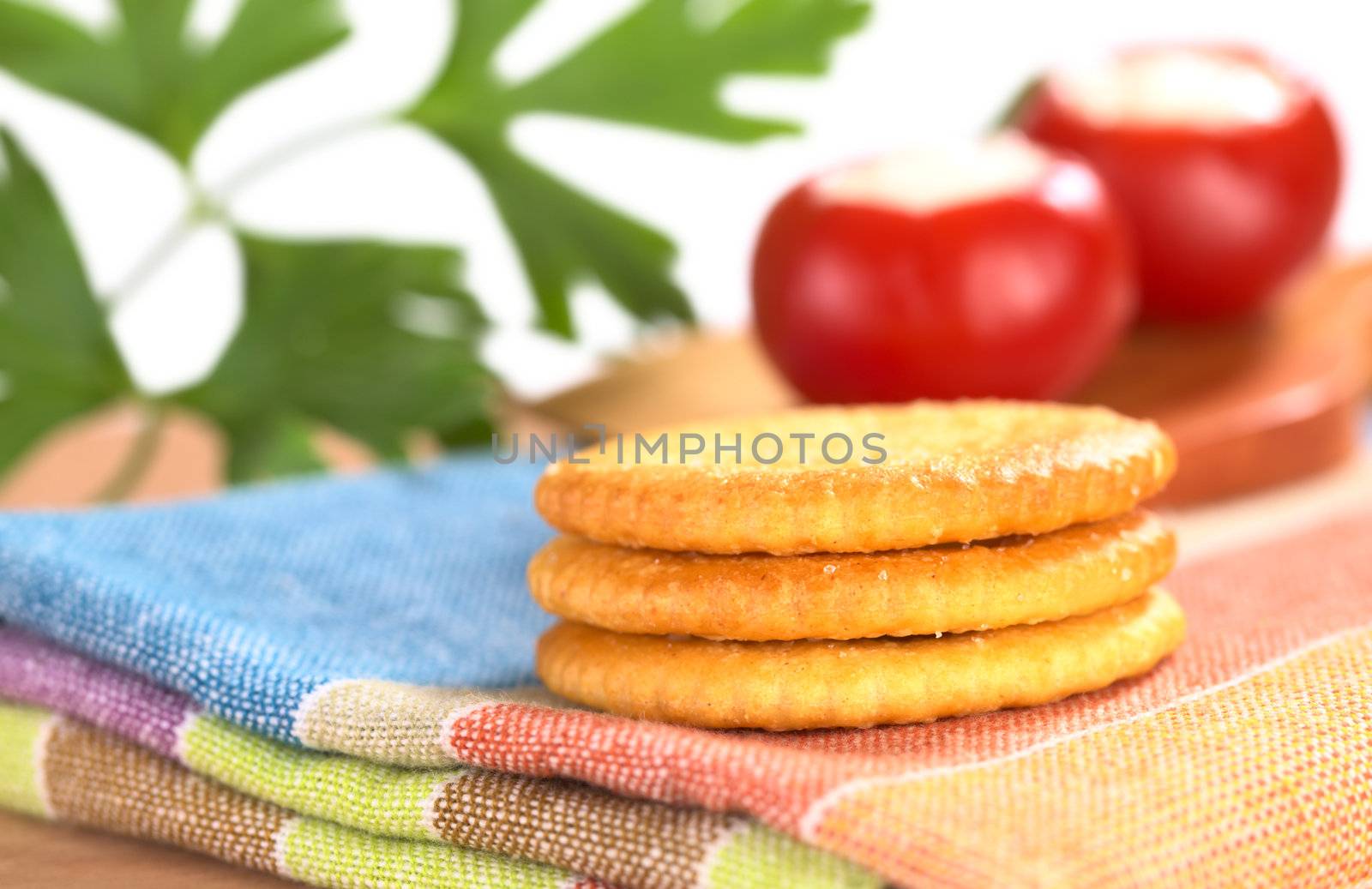 A pile of salty crackers on textile with round red pepper and parsley leaf in the back (Very Shallow Depth of Field, Focus on the front of the crackers)