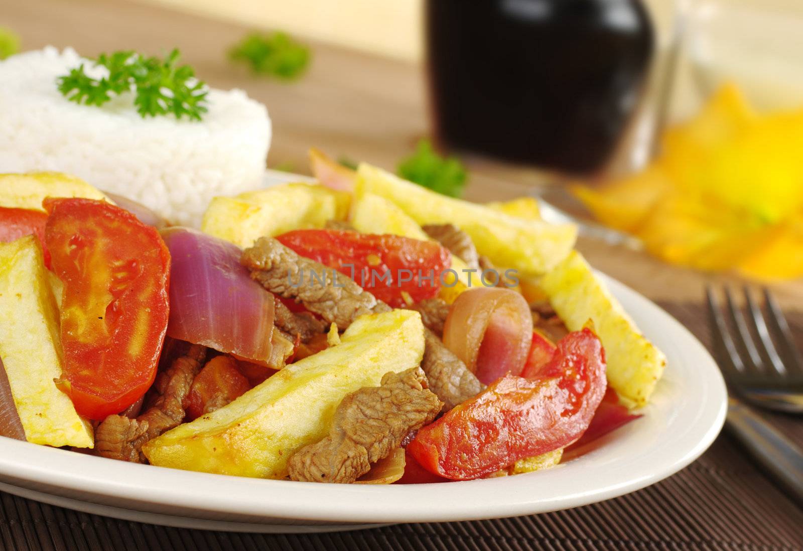 A typical Peruvian dish called Lomo Saltado which is made of beef, onions, tomatoes and is accompanied by fried potatoes and rice with cutlery and wine in the background (Selective Focus, Focus on the front of the dish)