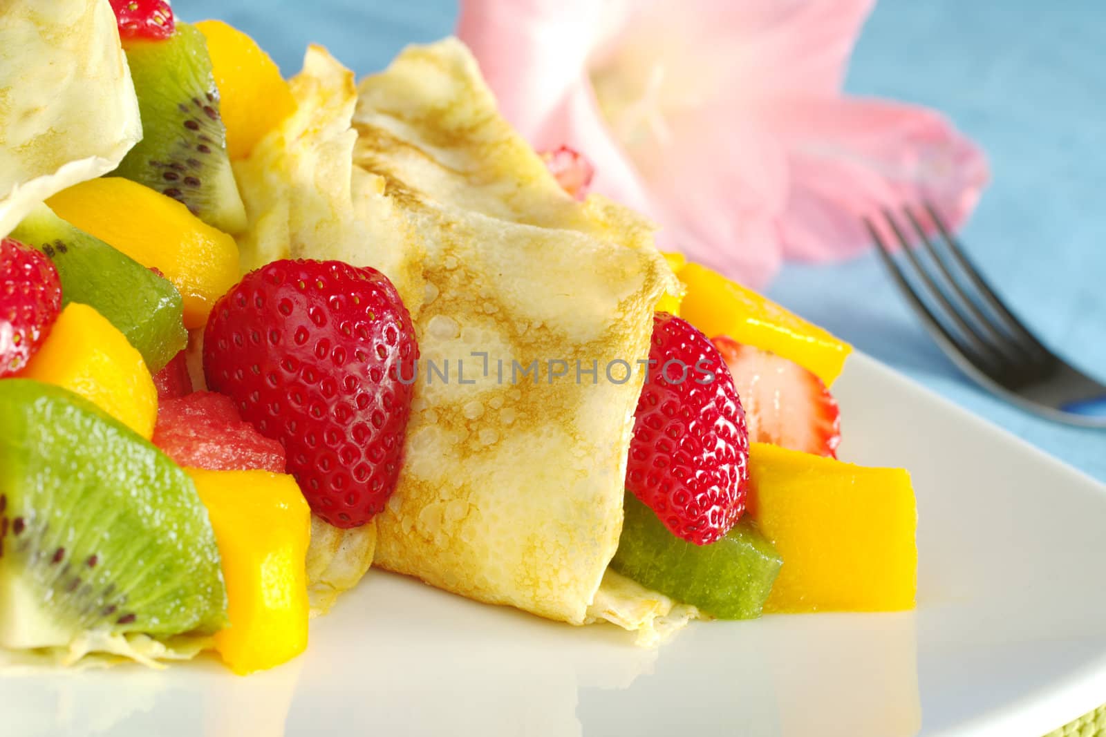Crepes filled with fresh fruits (strawberry, kiwi, mango, watermelon) with a pink gladiolus and a fork in the background (Selective Focus, Focus on the strawberry half and the crepe)