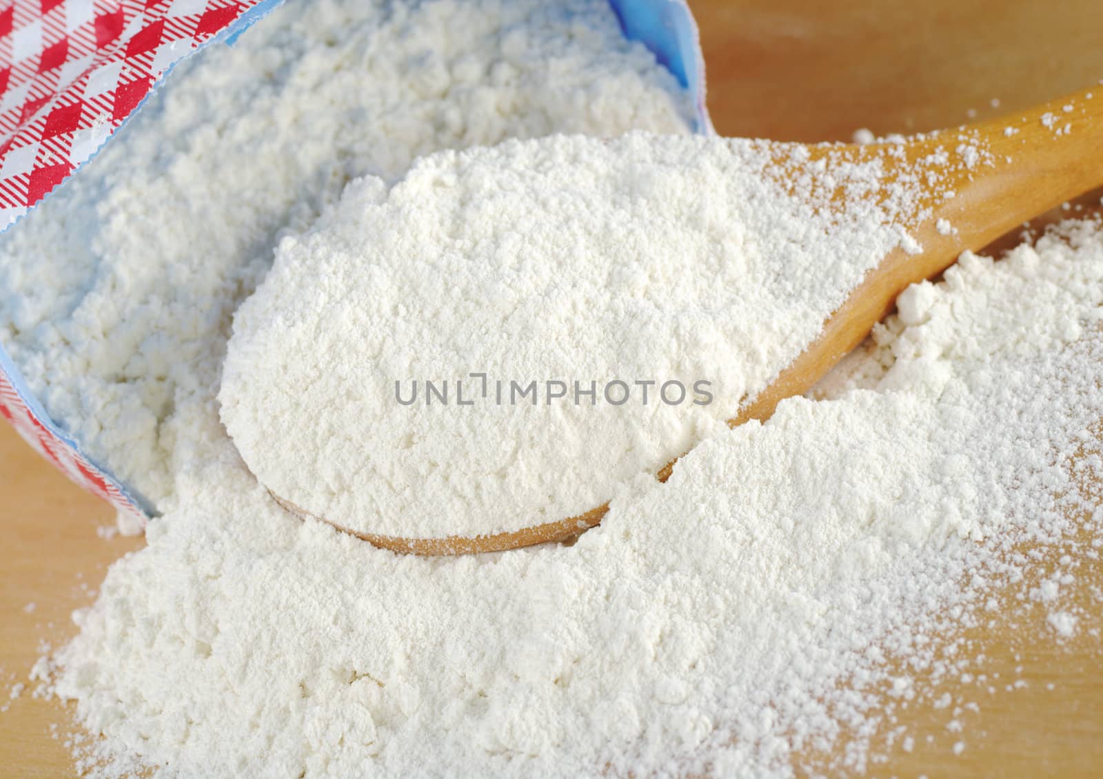 Wheat flour on wooden spoon lying in front of the paper package (Selective Focus, Focus on the flour on the spoon)