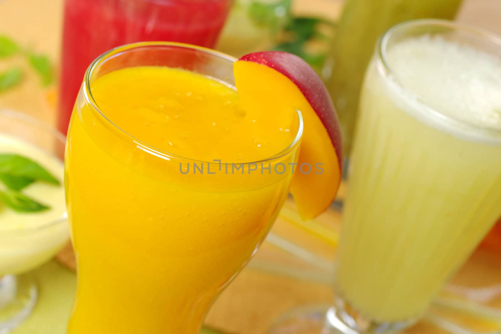 Fresh mango smoothie surrounded by watermelon, kiwi and pineapple smoothies and a cream cheese dessert on the left (Selective Focus, Focus on the front rim of the mango smoothie)