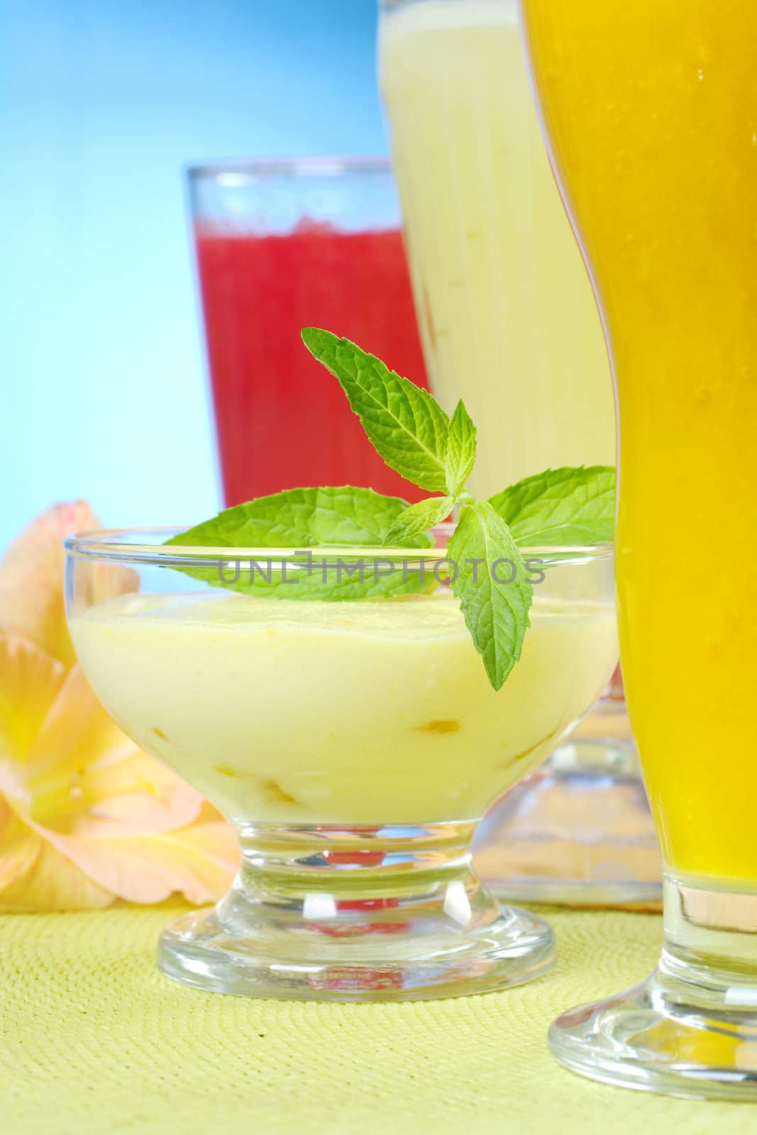 Light summer cream-cheese-mango dessert garnished with mint leaves with mango, pineapple and watermelon smoothies and gladiolus flower (Selective Focus, Focus on the mint leaf and the front of the bowl)