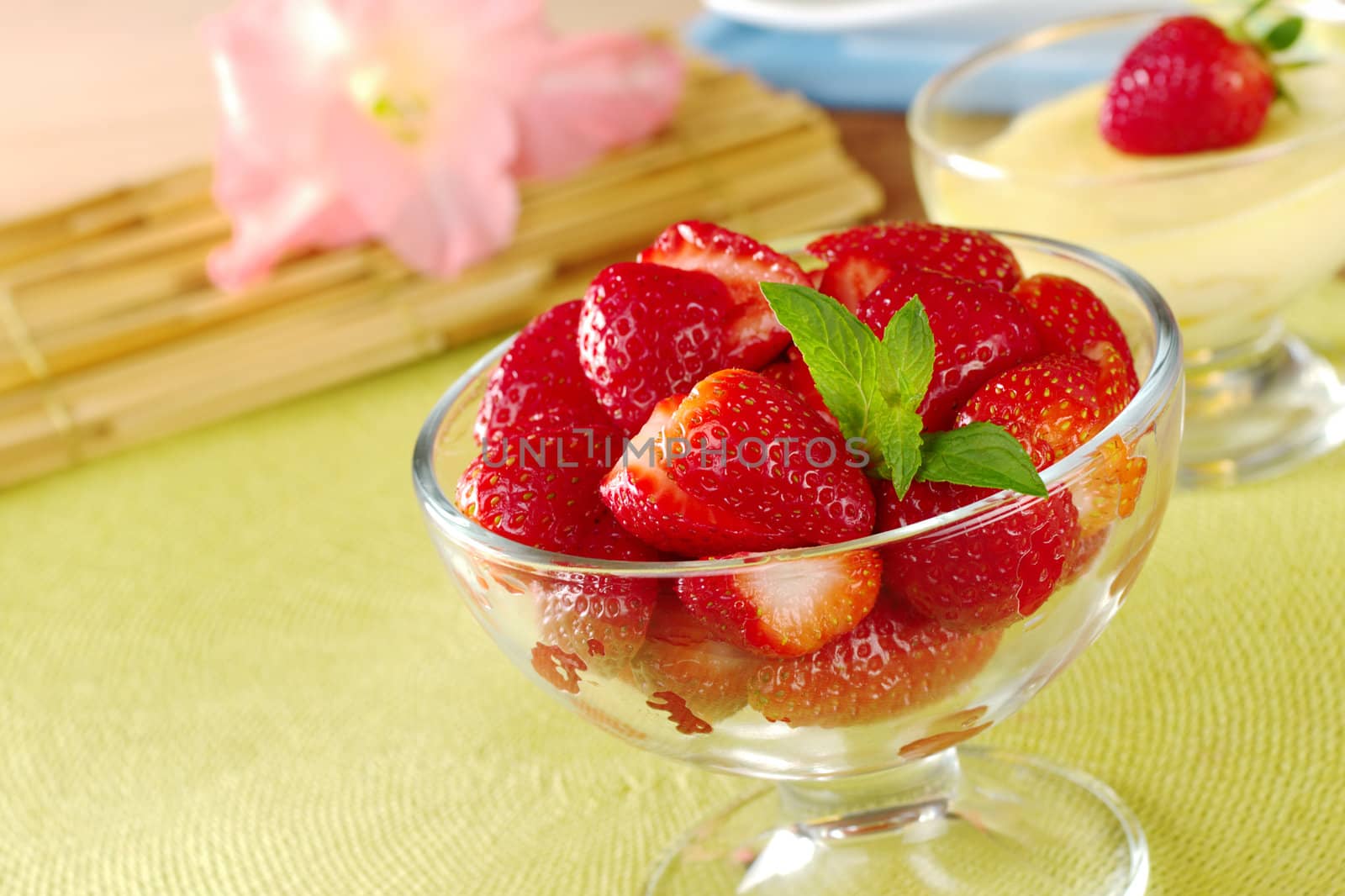 Fresh strawberries cut in half with mint leaf as garnish in a glass bowl  with a cream cheese dessert and gladiolus in the background (Selective Focus, Focus on the strawberries in the front and the mint leaf)
