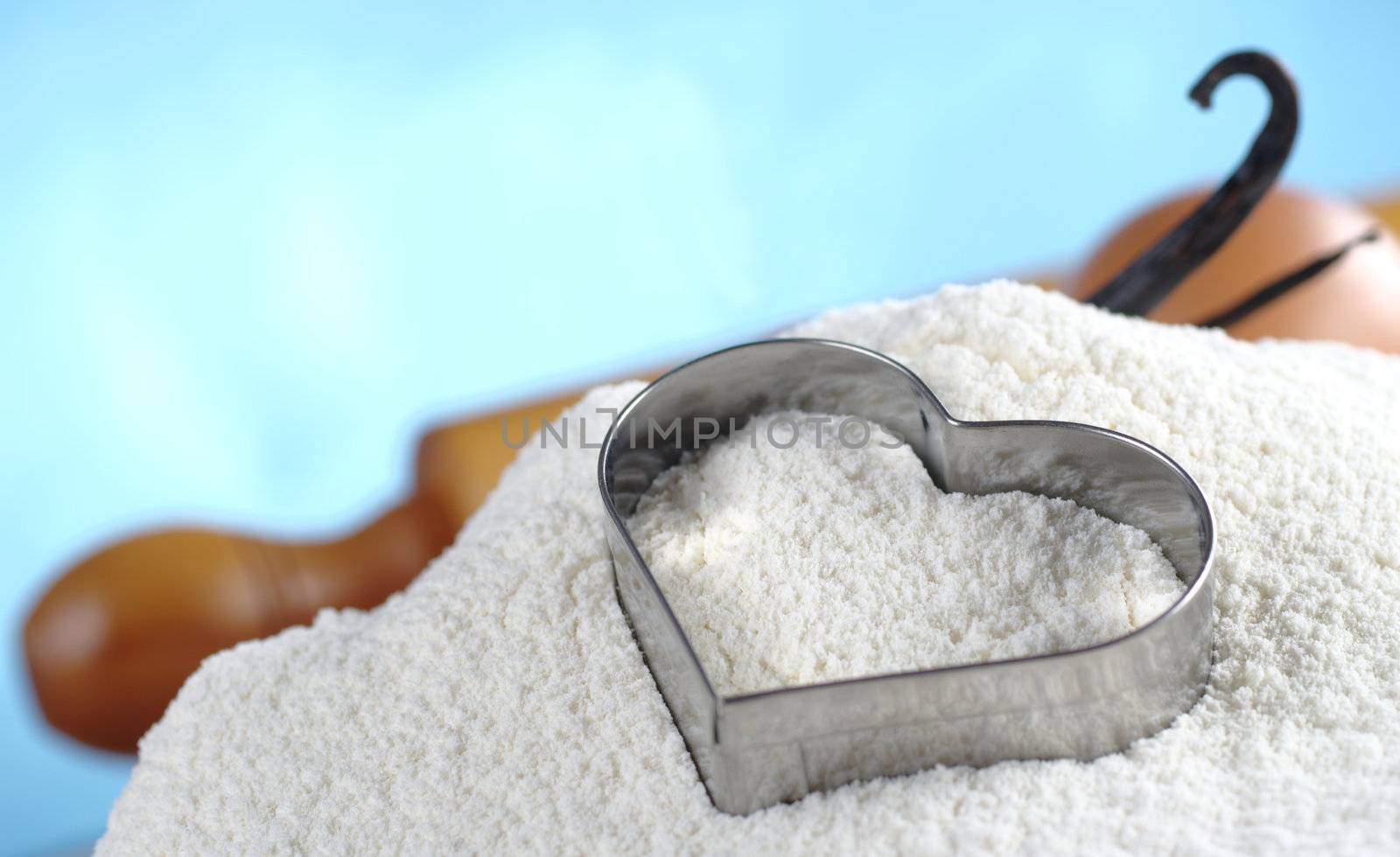 Heart shaped cookie cutter on flour with eggs, rolling pin and vanilla beans in the background (Selective Focus, Focus on the flour in the middle of the cookie cutter and parts of the cookie cutter)