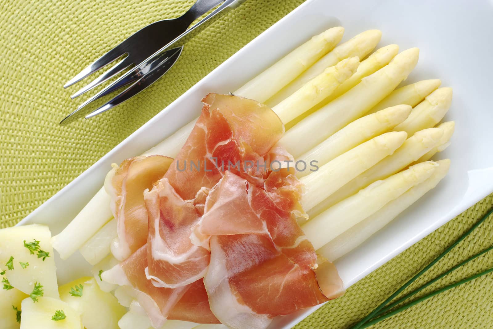 Fresh cooked white asparagus served with ham and potatoes on long plate with chives and cutlery on the sides