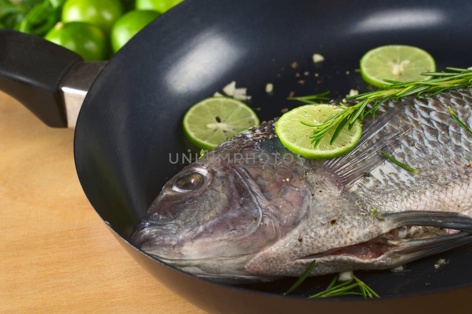 Raw tilapia with condiments (pepper corns, lime slices, garlic and rosemary) in frying pan (Selective Focus, Focus on the lime slice and rosemary on the fish and the eye)