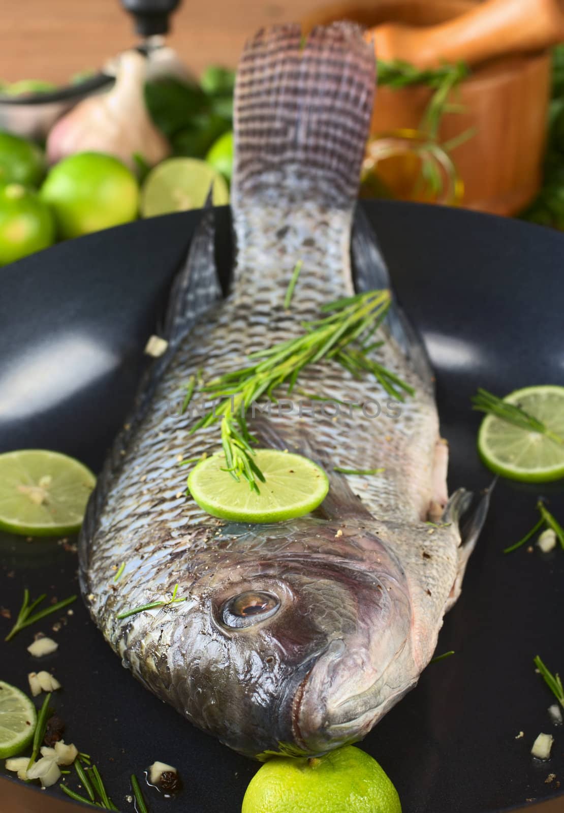 Raw tilapia with condiments (pepper corns, lime slices, garlic and rosemary) in frying pan (Selective Focus, Focus on the eye)