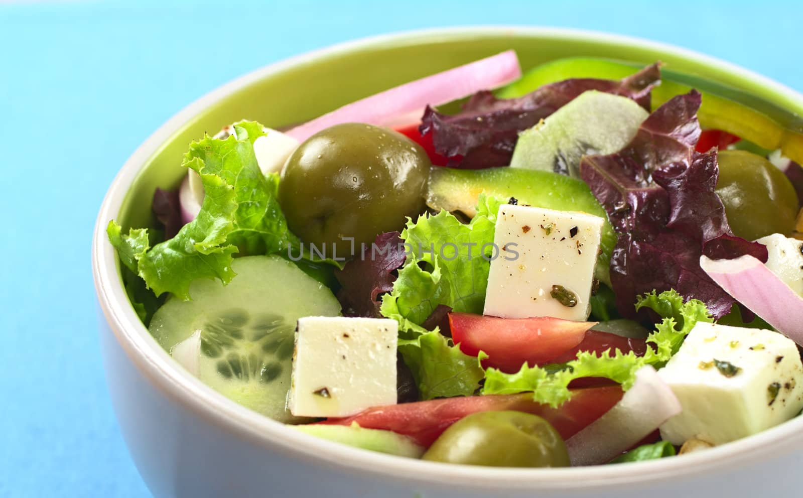 Greek salad out of cheese, green olives, tomato, green bell pepper, red onion, cucumber and lettuce in bowl on blue mat (Selective Focus, Focus on the cheese in the middle) 