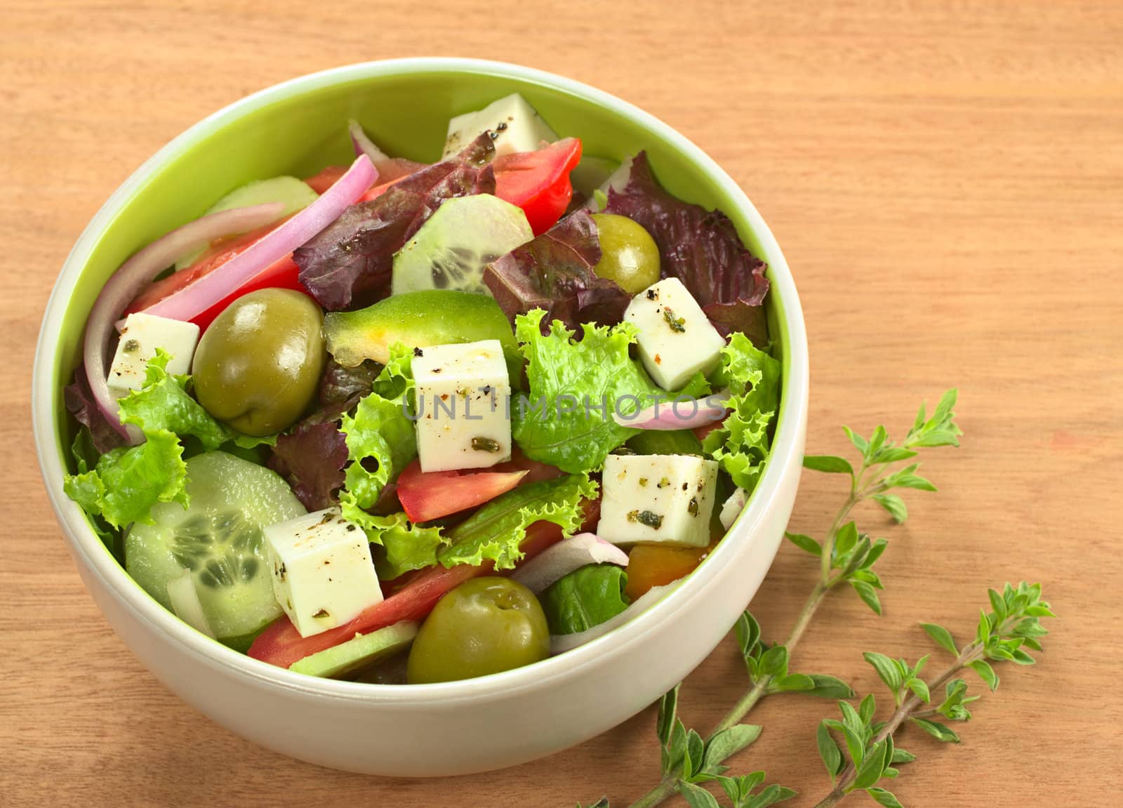 Greek salad out of cheese, green olives, tomato, green bell pepper, red onion, cucumber and lettuce with fresh oregano beside the bowl on wood (Selective Focus, Focus from the front to the middle of the bowl) 