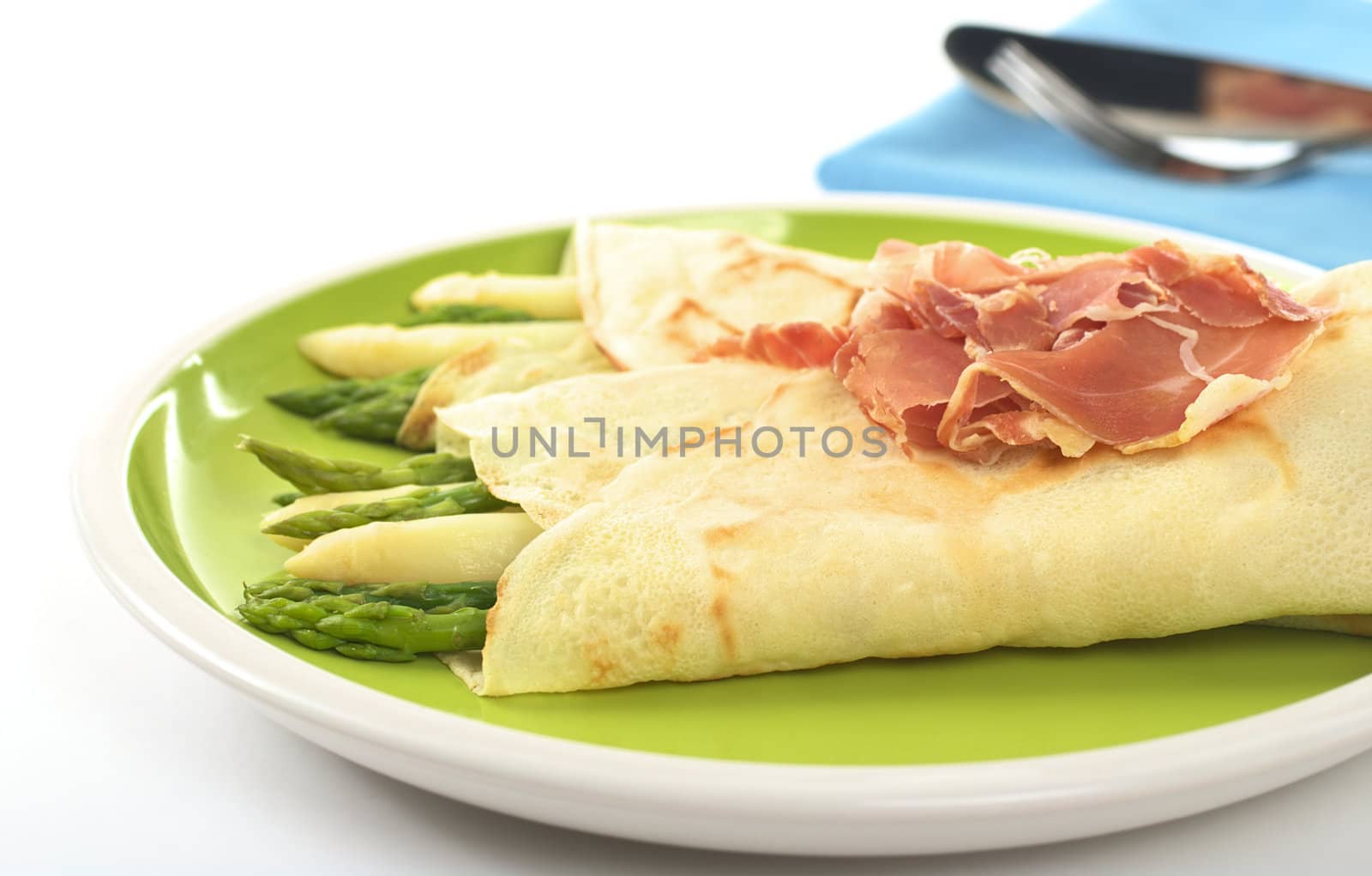 Green and white asparagus wrapped in crepes with ham as garnish on top (Selective Focus, Focus on the asparagus in the front and part of the ham)