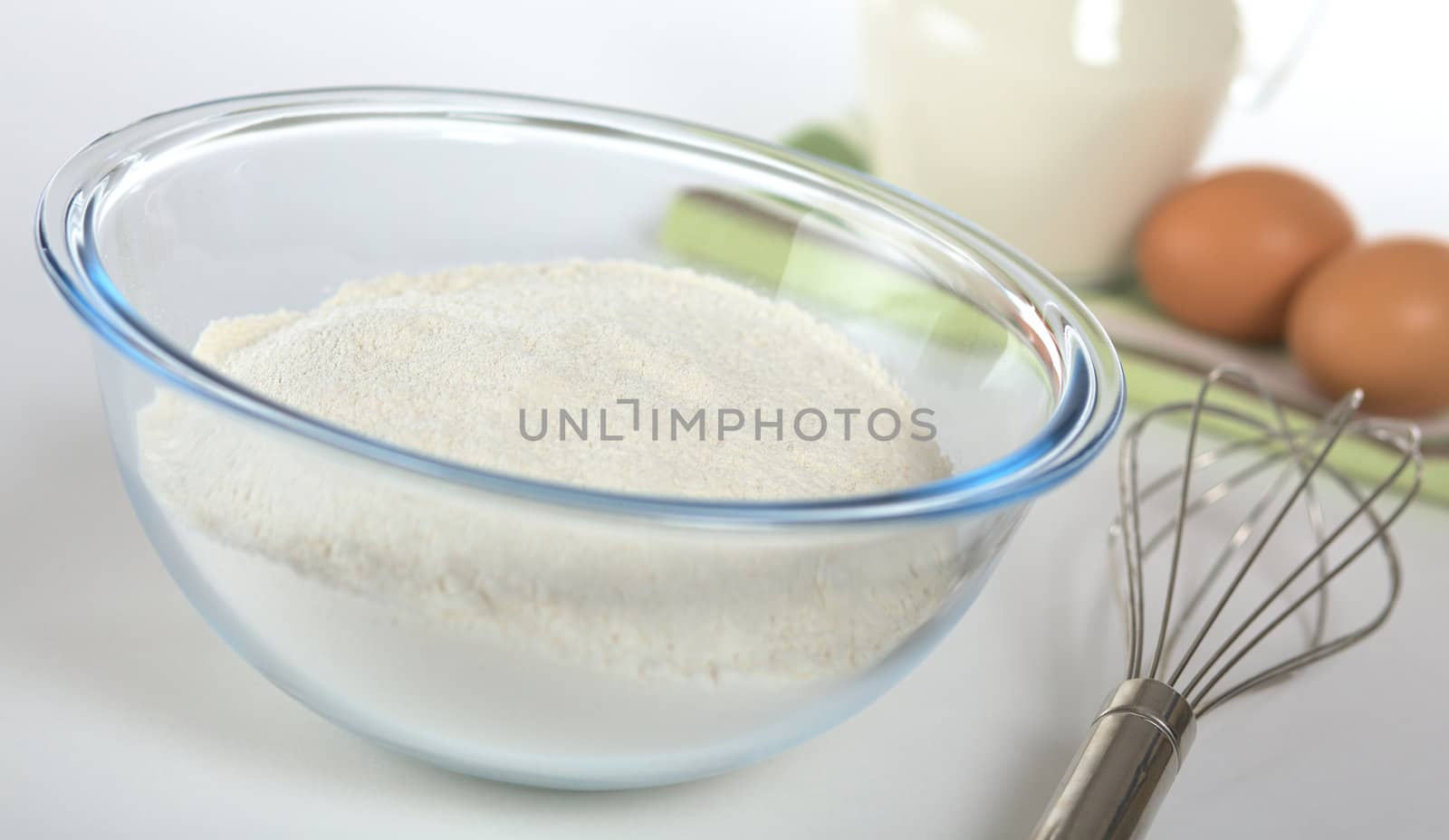 Preparing crepes with sieved flour in glass bowl and a metal beater on the side and milk and eggs in the back (Selective Focus, Focus on the middle of the flour in the bowl)