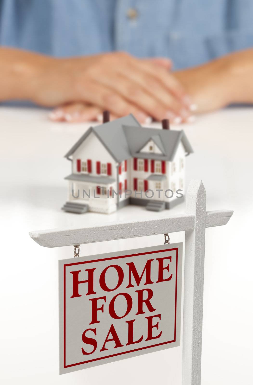 Womans Folded Hands Behind Model House and Home For Sale Real Estate Sign In Front on White Surface.