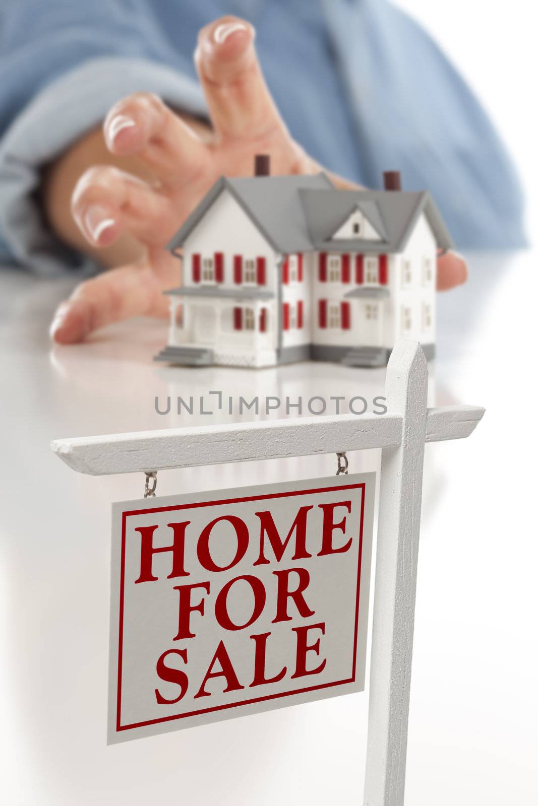 Real Estate Sign in Front of Womans Hand Reaching for Model House on a White Surface.