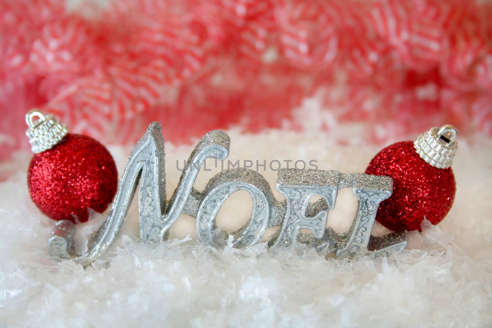 The word Noel made of glitter sitting in the middle of fake snow with two red christmas ornaments on each end.  Used selective focus and a shallow depth of field.