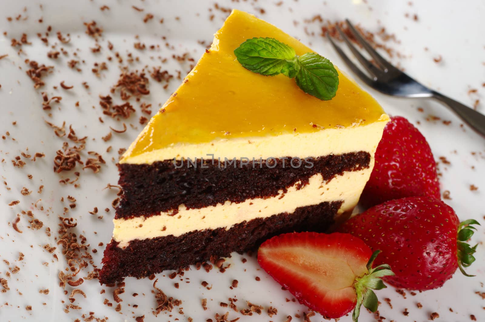 A piece of lucuma (South American fruit) cake garnished with mint leaf, chocolate shavings and strawberries served on a white plate with a pastry fork (Selective Focus, Focus on the mint leaf)