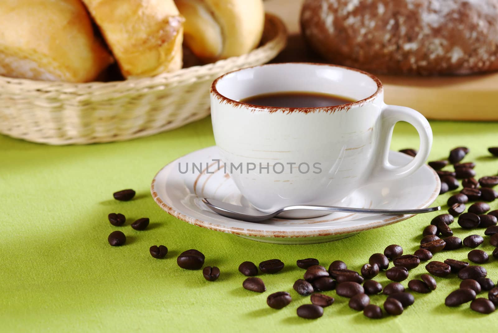 Coffee in cup surrounded by many coffee beans on green tablecloth with brown bread and rolls in the background (Selective Focus, Focus on the cup)