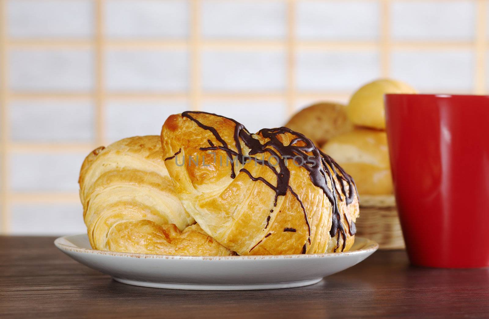 Two chocolate croissants with a red mug beside and bread in the background (Selective Focus, Focus on the front of the croissant) 