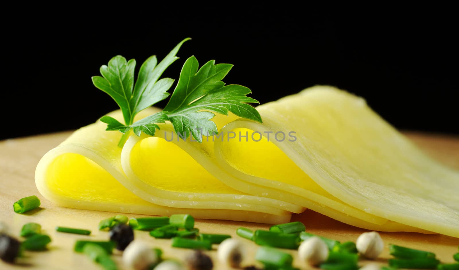 Cheese slices garnished with a parsley leaf and some shallot and pepper corns in the foreground on wooden board with black background (Selective Focus, Focus on the front of the slices and part of the parsley)