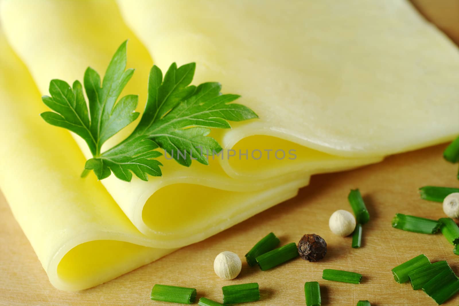 Cheese slices garnished with a parsley leaf and some shallot and pepper corns in the foreground on wooden board (Very Shallow Depth of Field, Focus on the upper front part of the cheese and part of the parsley)