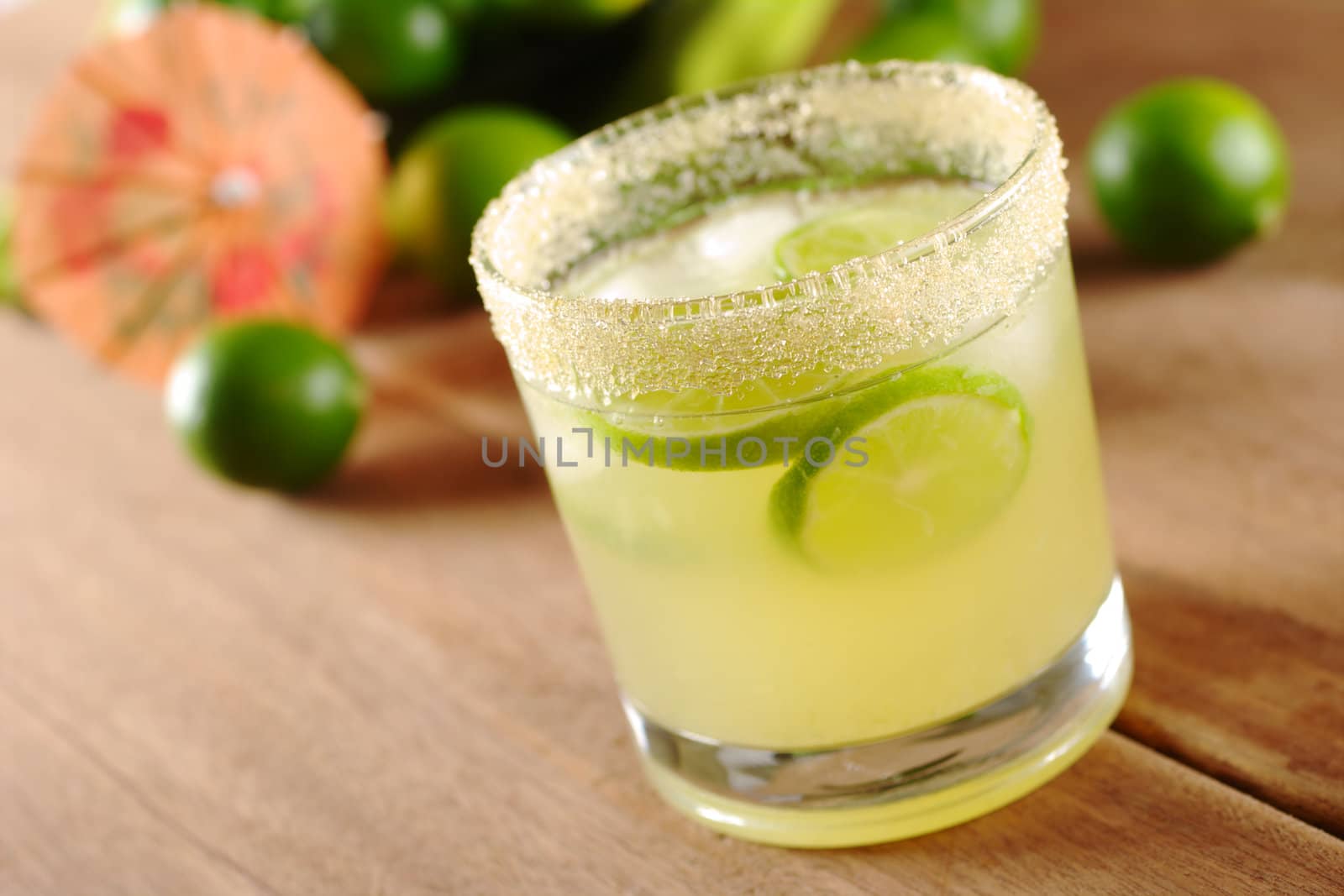 Fresh lemonade of green limes in sugar-rimmed glass on wooden board with limes and an orange paper sunshade in the background (Selective Focus, Focus on the front of the rim of the glass)