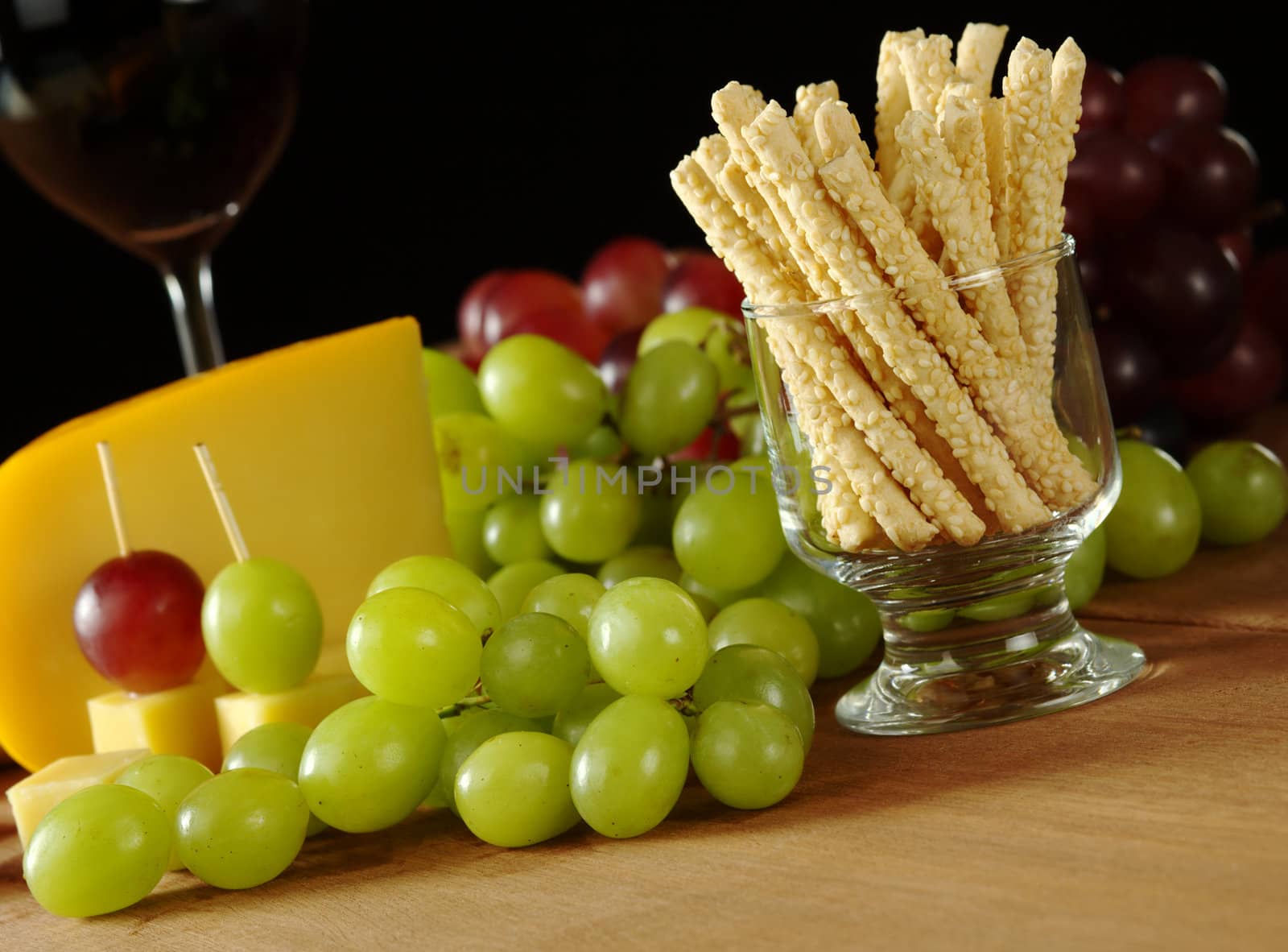 Snack: Sesame sticks with white grapes, some red grapes, cheese and red wine in glass in the background on wooden board (Selective Focus, Focus on the sesame sticks)